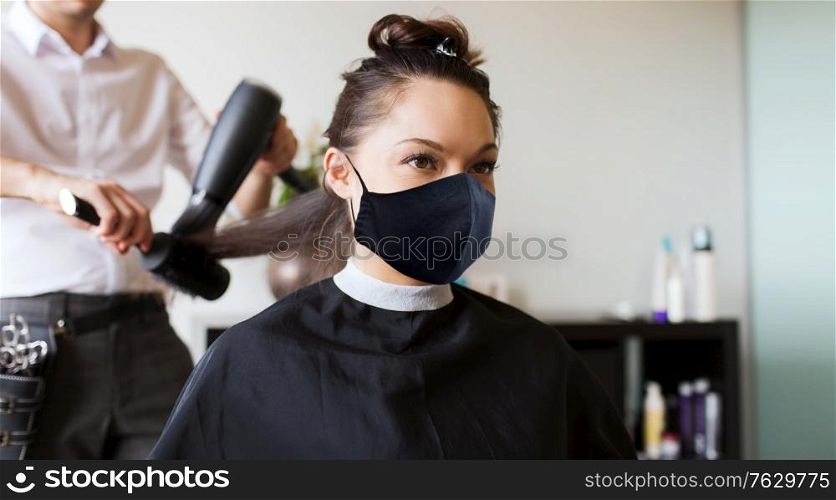 beauty, hairdressing and health safety concept - young woman wearing face protective medical mask for protection from virus disease and hairdresser with fan making hot styling at hair salon. happy woman with stylist making hairdo at salon