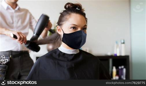 beauty, hairdressing and health safety concept - young woman wearing face protective medical mask for protection from virus disease and hairdresser with fan making hot styling at hair salon. happy woman with stylist making hairdo at salon