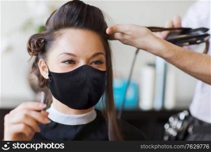 beauty, hairdressing and health safety concept - young woman wearing face protective medical mask for protection from virus disease and hairdresser with hair iron making hairdo at hair salon. happy woman with stylist making hairdo at salon