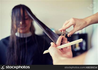beauty, hairstyle and people concept - happy young woman and hairdresser hands with scissors and comb cutting hair tips at salon