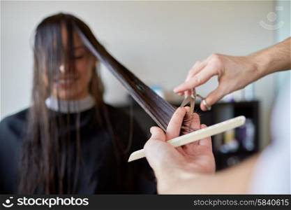 beauty, hairstyle and people concept - happy young woman and hairdresser hands with scissors and comb cutting hair tips at salon