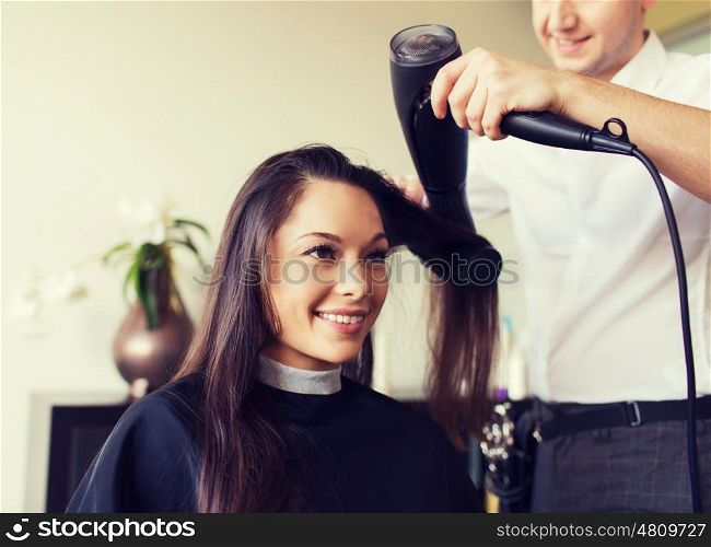 beauty, hairstyle and people concept - happy young woman and hairdresser with fan making hot styling at hair salon