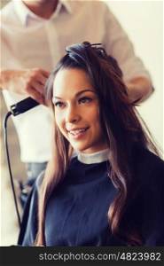 beauty, hairstyle and people concept - happy young woman with hairdresser curling hair and making hairdo at hair salon