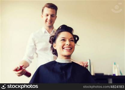 beauty, hairstyle and people concept - happy young woman with hairdresser with hair spray fixating hairdo at salon