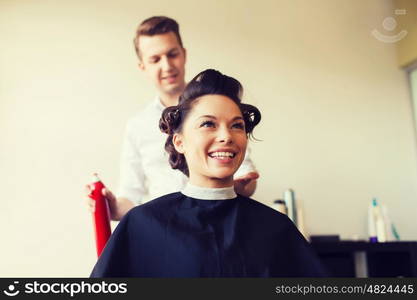 beauty, hairstyle and people concept - happy young woman with hairdresser with hair spray fixating hairdo at salon