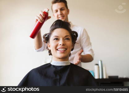 beauty, hairstyle and people concept - happy young woman with hairdresser with hair spray fixating hairdo at salon