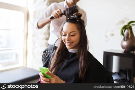 beauty, hairstyle and people concept - happy young woman with smartphone and hairdresser making hair styling at salon