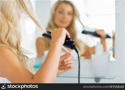 beauty, hairstyle, morning and people concept - close up of young woman with styling iron straightening her hair and looking to mirror at home bathroom