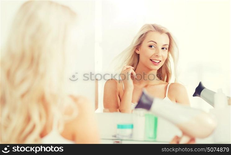 beauty, hairstyle, morning and people concept - smiling young woman with fan blow drying her hair looking to mirror at home bathroom. happy young woman with fan drying hair at bathroom