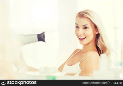beauty, hairstyle, morning and people concept - smiling young woman with fan blow drying her hair looking to mirror at home bathroom. happy young woman with fan drying hair at bathroom. happy young woman with fan drying hair at bathroom