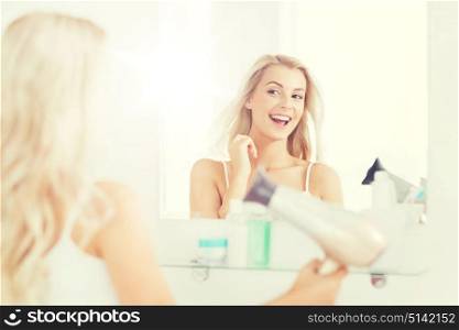 beauty, hairstyle, morning and people concept - smiling young woman with fan blow drying her hair looking to mirror at home bathroom. happy young woman with fan drying hair at bathroom