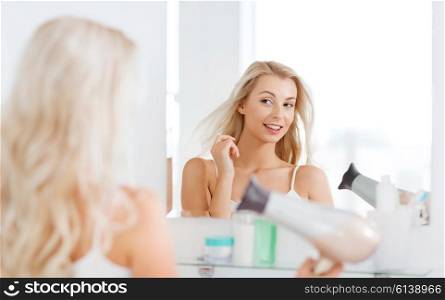 beauty, hairstyle, morning and people concept - smiling young woman with fan blow drying her hair looking to mirror at home bathroom