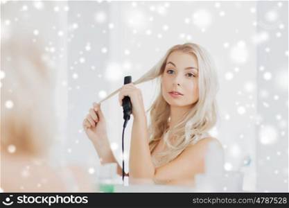 beauty, hairstyle, morning and people concept - smiling young woman with styling iron straightening her hair and looking to mirror at home bathroom over snow