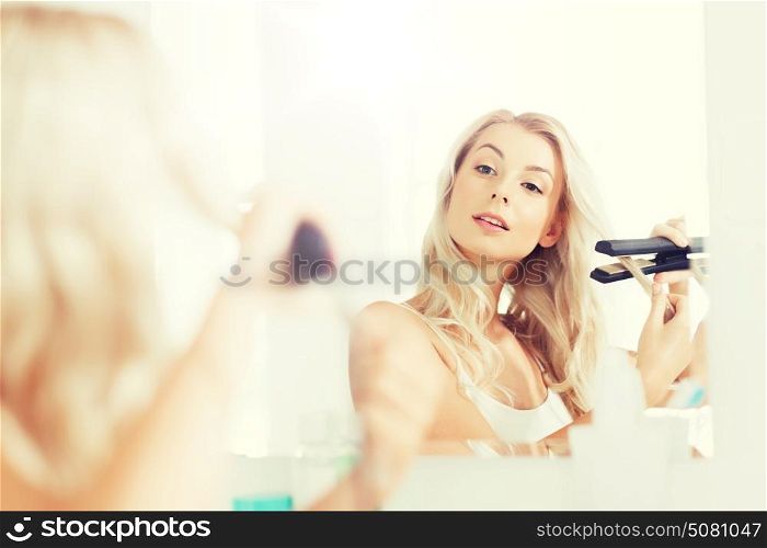 beauty, hairstyle, morning and people concept - smiling young woman with styling iron straightening her hair and looking to mirror at home bathroom. woman with styling iron doing her hair at bathroom