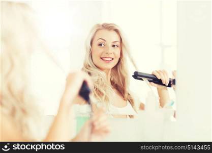 beauty, hairstyle, morning and people concept - smiling young woman with styling iron straightening her hair and looking to mirror at home bathroom. woman with styling iron doing her hair at bathroom