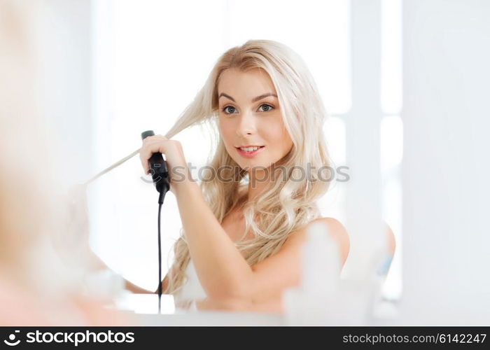 beauty, hairstyle, morning and people concept - smiling young woman with styling iron straightening her hair and looking to mirror at home bathroom