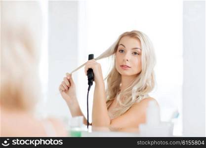 beauty, hairstyle, morning and people concept - smiling young woman with styling iron straightening her hair and looking to mirror at home bathroom