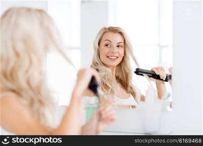beauty, hairstyle, morning and people concept - smiling young woman with styling iron straightening her hair and looking to mirror at home bathroom