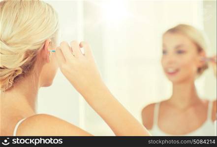 beauty, hygiene and people concept - close up of smiling young woman cleaning ear with cotton swab and looking to mirror at home bathroom. woman cleaning ear with cotton swab at bathroom. woman cleaning ear with cotton swab at bathroom