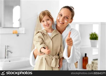beauty, hygiene and people concept - happy smiling mother and daughter with moisturizer on their faces showing thumbs up gesture in bathroom. mother and daughter showing thumbs up in bathroom