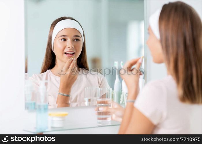 beauty, hygiene and people concept - happy teenage girl with cod liver oil capsule and glass of water looking in mirror at bathroom. teenage girl taking cod liver oil at mirror