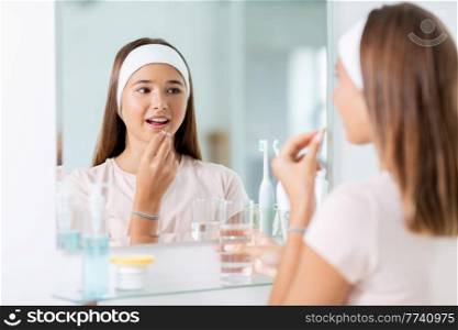 beauty, hygiene and people concept - happy teenage girl with cod liver oil capsule and glass of water looking in mirror at bathroom. teenage girl taking cod liver oil at mirror