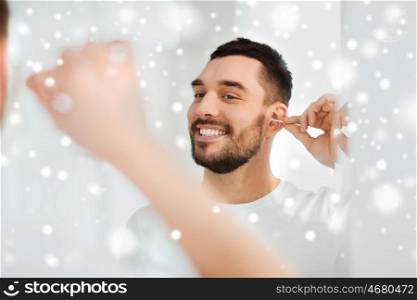 beauty, hygiene and people concept - smiling young man cleaning ear with cotton swab and looking to mirror at home bathroom over snow