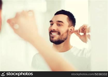 beauty, hygiene and people concept - smiling young man cleaning ear with cotton swab and looking to mirror at home bathroom. man cleaning ear with cotton swab at bathroom