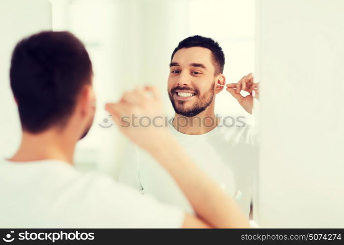 beauty, hygiene and people concept - smiling young man cleaning ear with cotton swab and looking to mirror at home bathroom. man cleaning ear with cotton swab at bathroom