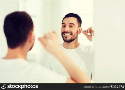 beauty, hygiene and people concept - smiling young man cleaning ear with cotton swab and looking to mirror at home bathroom. man cleaning ear with cotton swab at bathroom