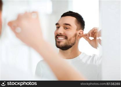 beauty, hygiene and people concept - smiling young man cleaning ear with cotton swab and looking to mirror at home bathroom