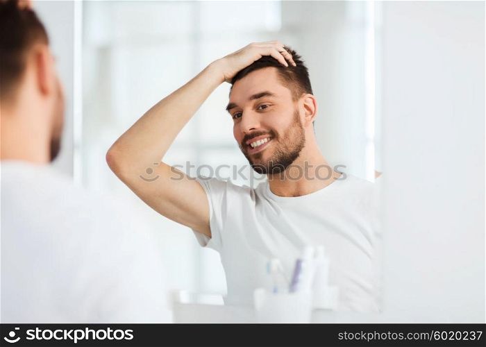 beauty, hygiene and people concept - smiling young man looking to mirror and styling hair at home bathroom