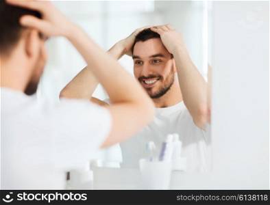 beauty, hygiene and people concept - smiling young man looking to mirror and styling hair at home bathroom