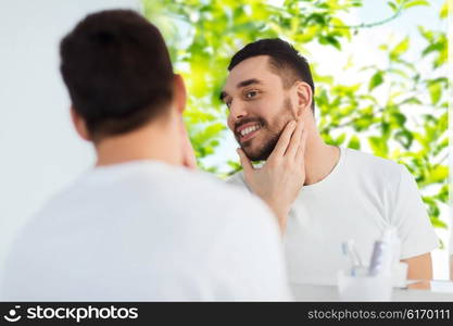 beauty, hygiene and people concept - smiling young man looking to mirror at home bathroom over green natural background