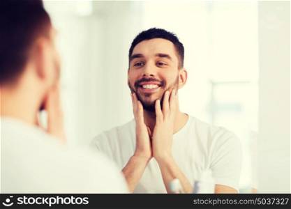 beauty, hygiene and people concept - smiling young man looking to mirror at home bathroom. happy young man looking to mirror at home bathroom