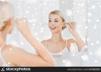 beauty, hygiene and people concept - smiling young woman cleaning ear with cotton swab and looking to mirror at home bathroom over snow