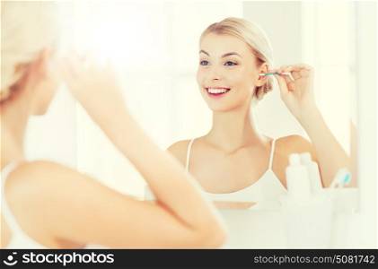 beauty, hygiene and people concept - smiling young woman cleaning ear with cotton swab and looking to mirror at home bathroom. woman cleaning ear with cotton swab at bathroom