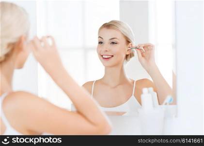 beauty, hygiene and people concept - smiling young woman cleaning ear with cotton swab and looking to mirror at home bathroom