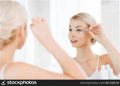 beauty, hygiene and people concept - smiling young woman fixing makeup with cotton swab and looking to mirror at home bathroom