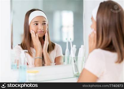 beauty, hygiene and people concept - teenage girl touching her face and looking in mirror at bathroom. teenage girl looking in mirror at bathroom