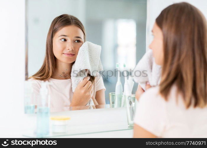beauty, hygiene and people concept - teenage girl wiping her face with towel and looking in mirror at bathroom. teenage girl wiping face with towel in bathroom
