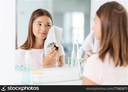 beauty, hygiene and people concept - teenage girl wiping her face with towel and looking in mirror at bathroom. teenage girl wiping face with towel in bathroom