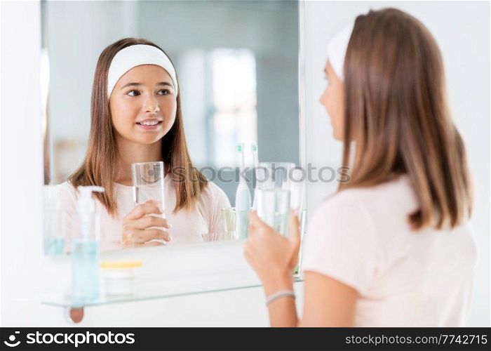 beauty, hygiene and people concept - teenage girl with glass of water looking in mirror at bathroom. teenage girl with glass of water looking in mirror