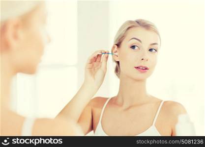 beauty, hygiene and people concept - young woman cleaning ear with cotton swab and looking to mirror at home bathroom. woman cleaning ear with cotton swab at bathroom