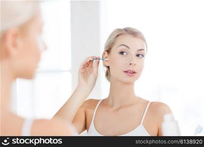 beauty, hygiene and people concept - young woman cleaning ear with cotton swab and looking to mirror at home bathroom