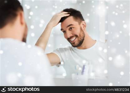 beauty, hygiene, hairstyle and people concept - smiling young man looking to mirror and styling hair at home bathroom over snow
