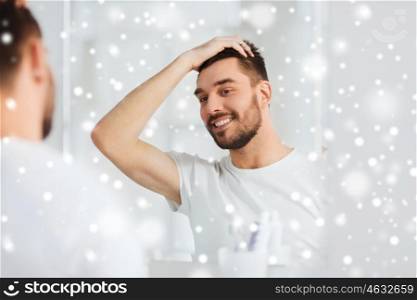 beauty, hygiene, hairstyle and people concept - smiling young man looking to mirror and styling hair at home bathroom over snow
