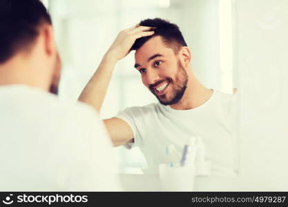 beauty, hygiene, hairstyle and people concept - smiling young man looking to mirror and styling hair at home bathroom. happy young man looking to mirror at home bathroom