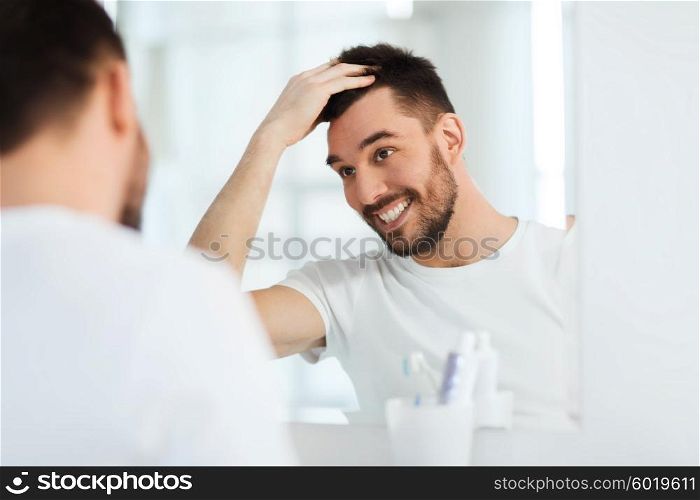 beauty, hygiene, hairstyle and people concept - smiling young man looking to mirror and styling hair at home bathroom