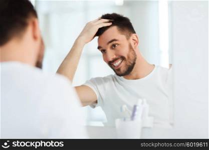 beauty, hygiene, hairstyle and people concept - smiling young man looking to mirror and styling hair at home bathroom